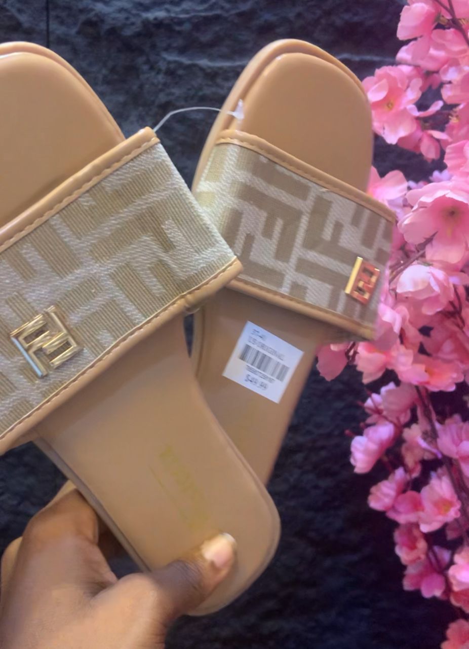 Beige slide sandals with a brand logo held by a hand, pink flowers in the background