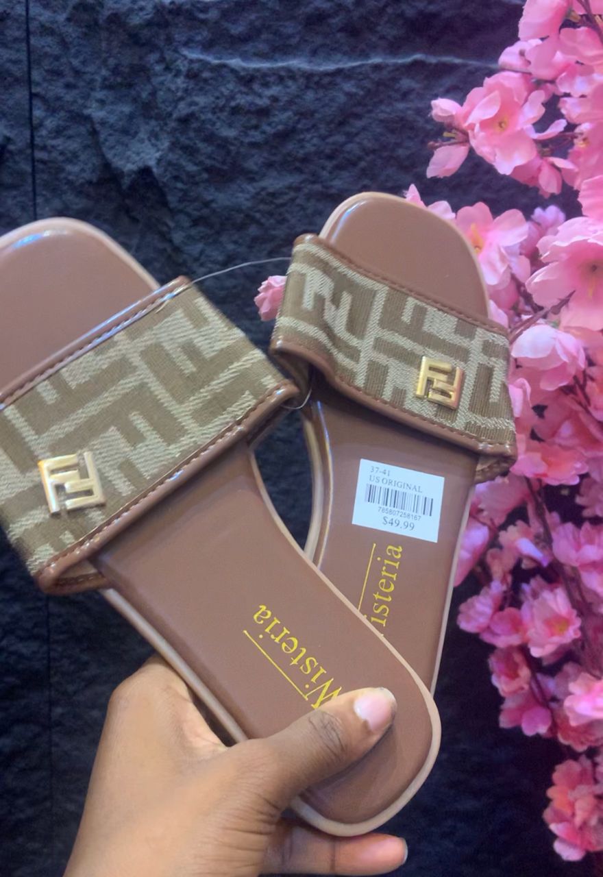 Pair of brown slides with patterned straps held by a hand against a dark background with pink flowers.